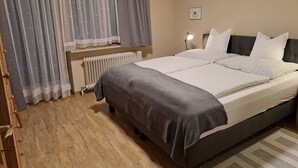 Una cama bien hecha con ropa de cama blanca y gris, una lámpara de noche y una ventana con cortinas translúcidas.