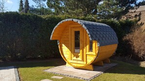 A wooden barrel-shaped structure with a door, surrounded by a garden and trees. - Holiday house of high quality and cozy, sauna, 150m to the beach, quiet natural location (Rankwitz (OT Quilitz))