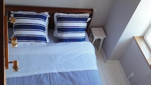 A bedroom with a wooden bed, blue and white checkered bedspread, two pillows, a nightstand, a wall-mounted painting, and a window with a view.