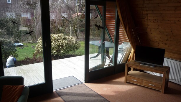 A room with a large sliding glass door leading to a patio with a table and chairs, a wooden cabinet, and a television. - Family-friendly holiday house with large garden and great features (Butjadingen-Burhave)