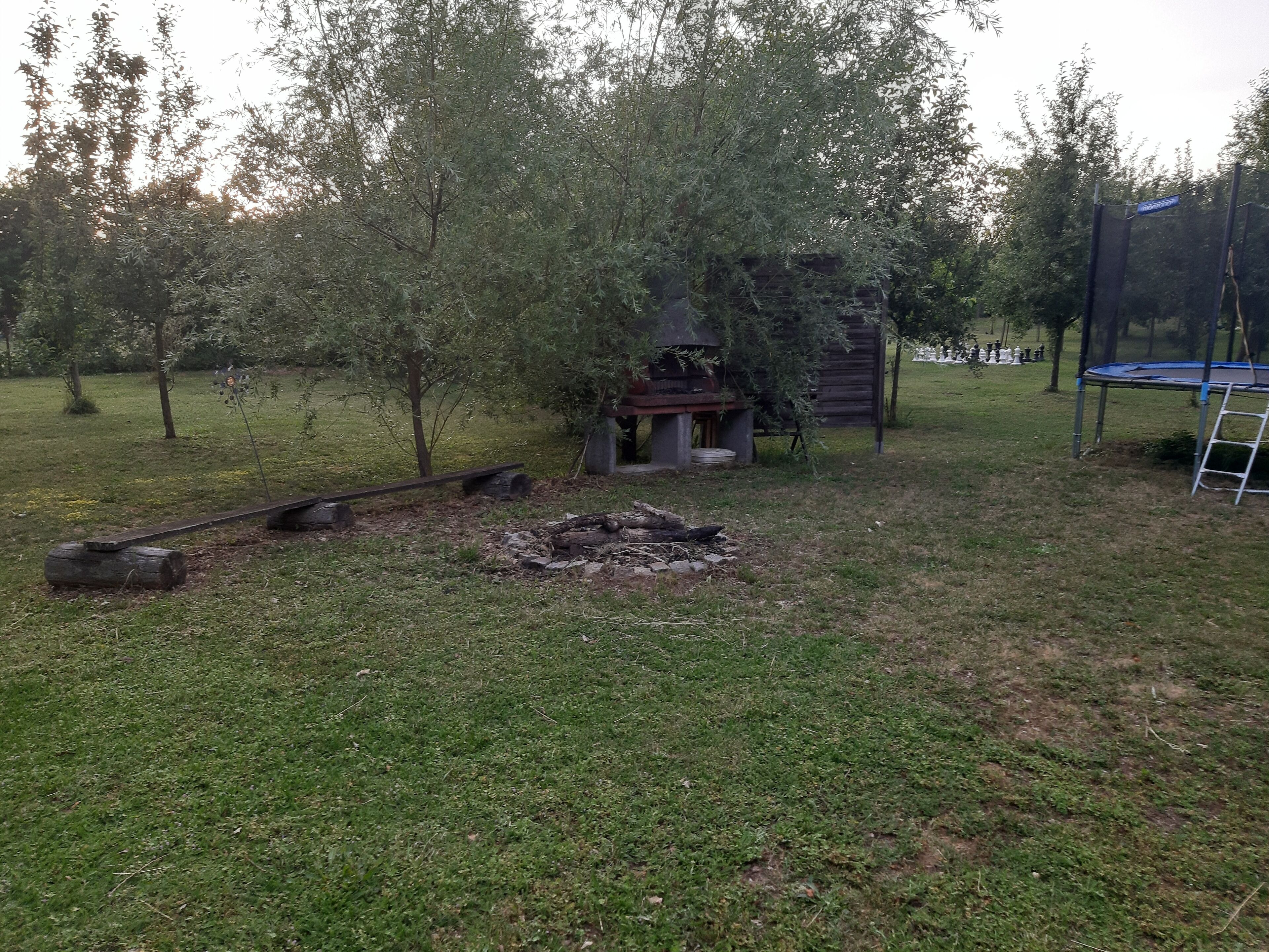 Ein grünes Außenbereich mit einer kleinen Feuerstelle, einer Holzkonstruktion und einem Trampolin.