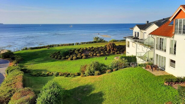Una proprietà costiera con un ampio prato, vista sul mare e un edificio moderno con numerose finestre e un tetto rosso.