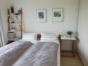 Une chambre avec deux lits, une table de chevet avec une lampe, une étagère avec des livres et des plantes, et deux tableaux encadrés accrochés au mur.
