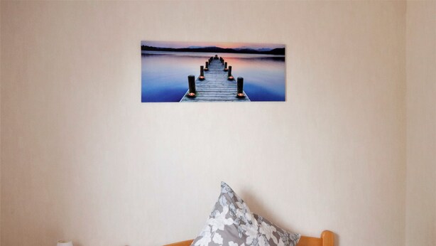 A bedroom with a wooden bed, a nightstand with a lamp, and a wall-mounted picture of a pier. - Holiday in a quiet location (Nideggen)