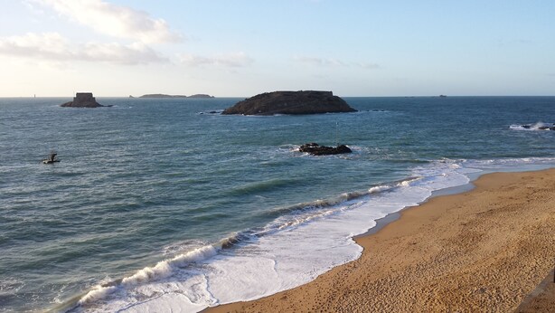 A beach with calm waves, distant rocky islands, and a clear sky.