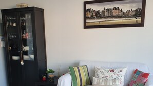A living room with a white sofa, a black cabinet, a framed cityscape picture, and a white coffee table with a woven placemat.