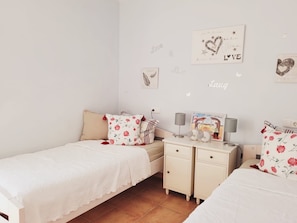 Une chambre avec deux lits simples, une table de chevet avec une lampe et des éléments de décoration murale, notamment un panneau en forme de cœur et une illustration d’oiseau.