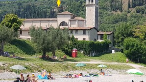 A building with a bell tower, surrounded by greenery and a beach area with people relaxing. - Modern apartment with Seepanoramablick in a quiet area with large infinity pool (Toscolano Maderno)
