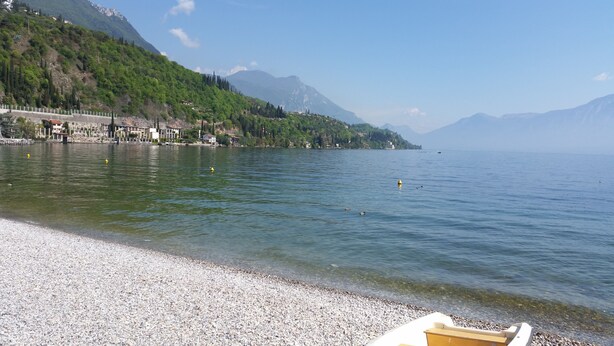 A lake with a pebbled shore, a row of buildings along the water's edge, and mountains in the background. - Modern apartment with Seepanoramablick in a quiet area with large infinity pool (Toscolano Maderno)