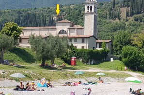 A building with a bell tower, surrounded by greenery and a beach area with people relaxing. - Modern apartment with Seepanoramablick in a quiet area with large infinity pool (Toscolano Maderno)