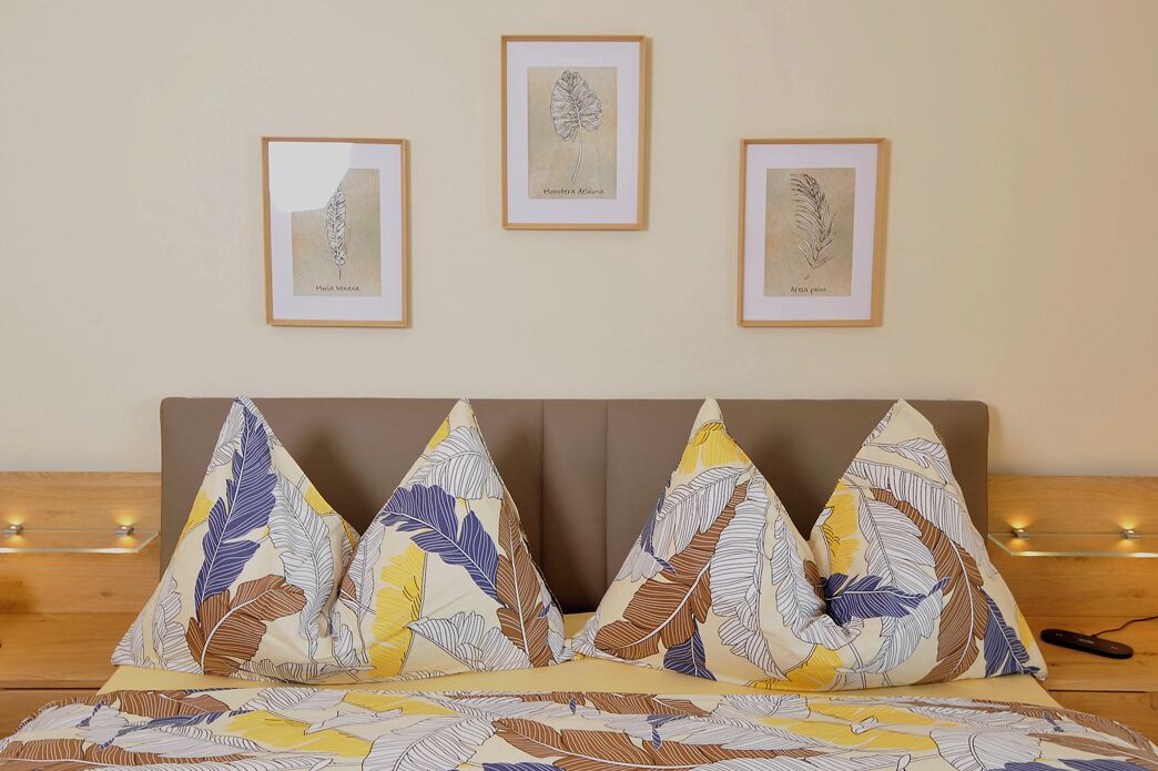 A bed with leaf-patterned pillows, a framed botanical illustration on the wall, and a wooden bedside table with a remote control.