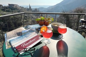 A table with a magazine, a bowl of fruit, and two glasses of red drink on a balcony with a scenic view.