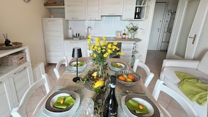 Ein Essbereich mit einem Tisch, gedeckt für vier Personen, umgeben von weißen Stühlen. Auf dem Tisch steht eine Vase mit gelben Blumen, und es gibt Teller mit grünen Servietten. Im Hintergrund sind ein Kühlschrank und Küchenschränke zu sehen.