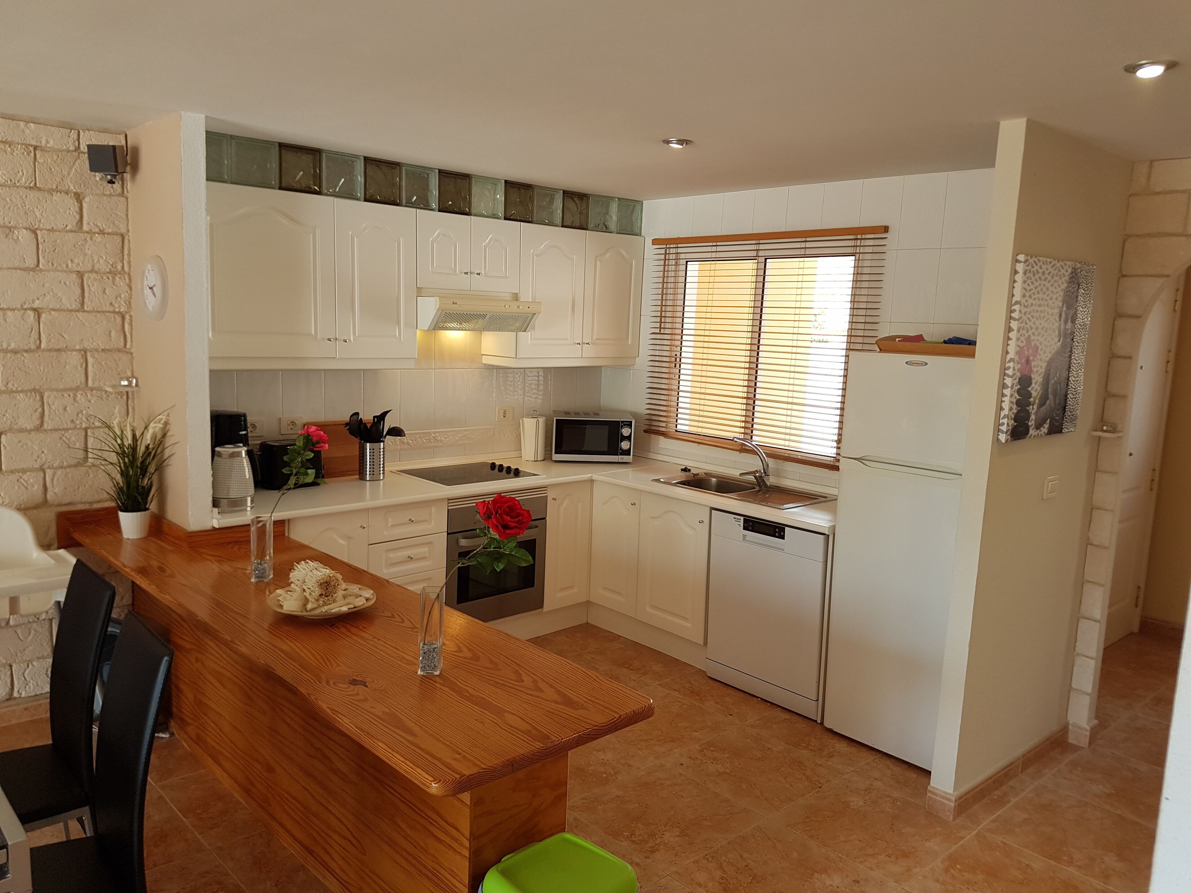 A compact kitchen with white cabinets, a wooden dining table, a green trash bin, and a window with blinds.