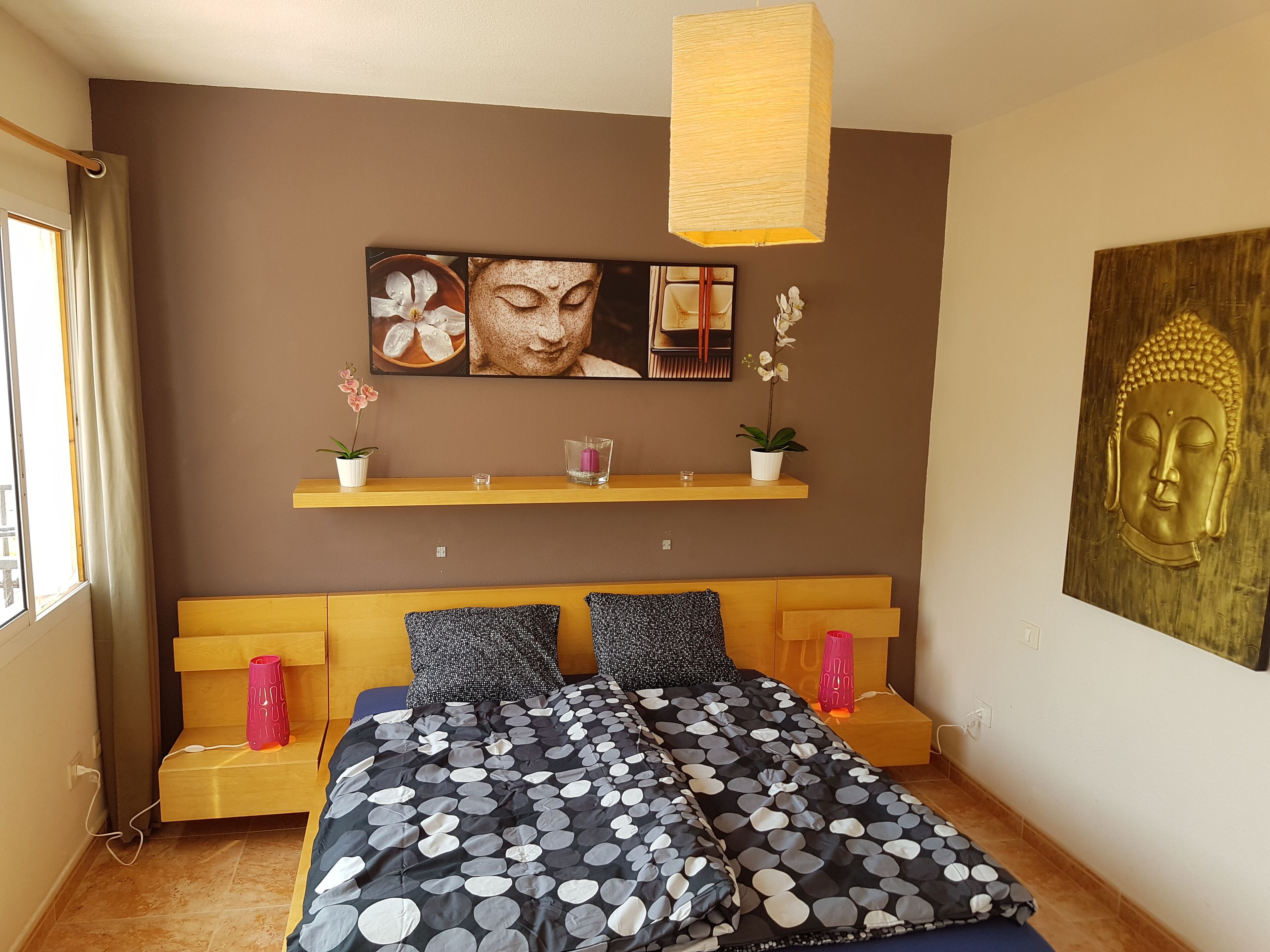 Un dormitorio con cama, cabecero de madera, repisa con adornos, estatua de Buda y ventana con cortinas.