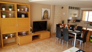 A modern living room with a TV, wooden entertainment unit, dining area with chairs and table, kitchen with appliances, and decorative plants. - Villa Roque del Conde 3 with fantastic sea view, Terrace, Pool, Wifi, SAT-TV (Costa Adeje)