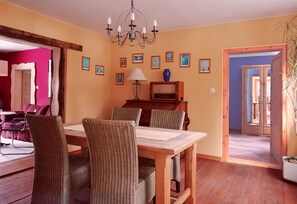 Ein Essbereich mit Holztisch und Stühlen, einer Chandelierleuchtre, gerahmten Bildern an der Wand und Blick in einen roten Raum durch eine offene Tür.