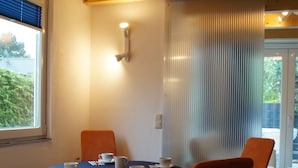A dining area with a table set for four, surrounded by four orange chairs. A window with blinds is visible to the left, and a door with a glass panel is to the right.