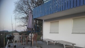 Une terrasse avec des meubles d’extérieur, un barbecue et un bâtiment bleu avec des volets blancs.