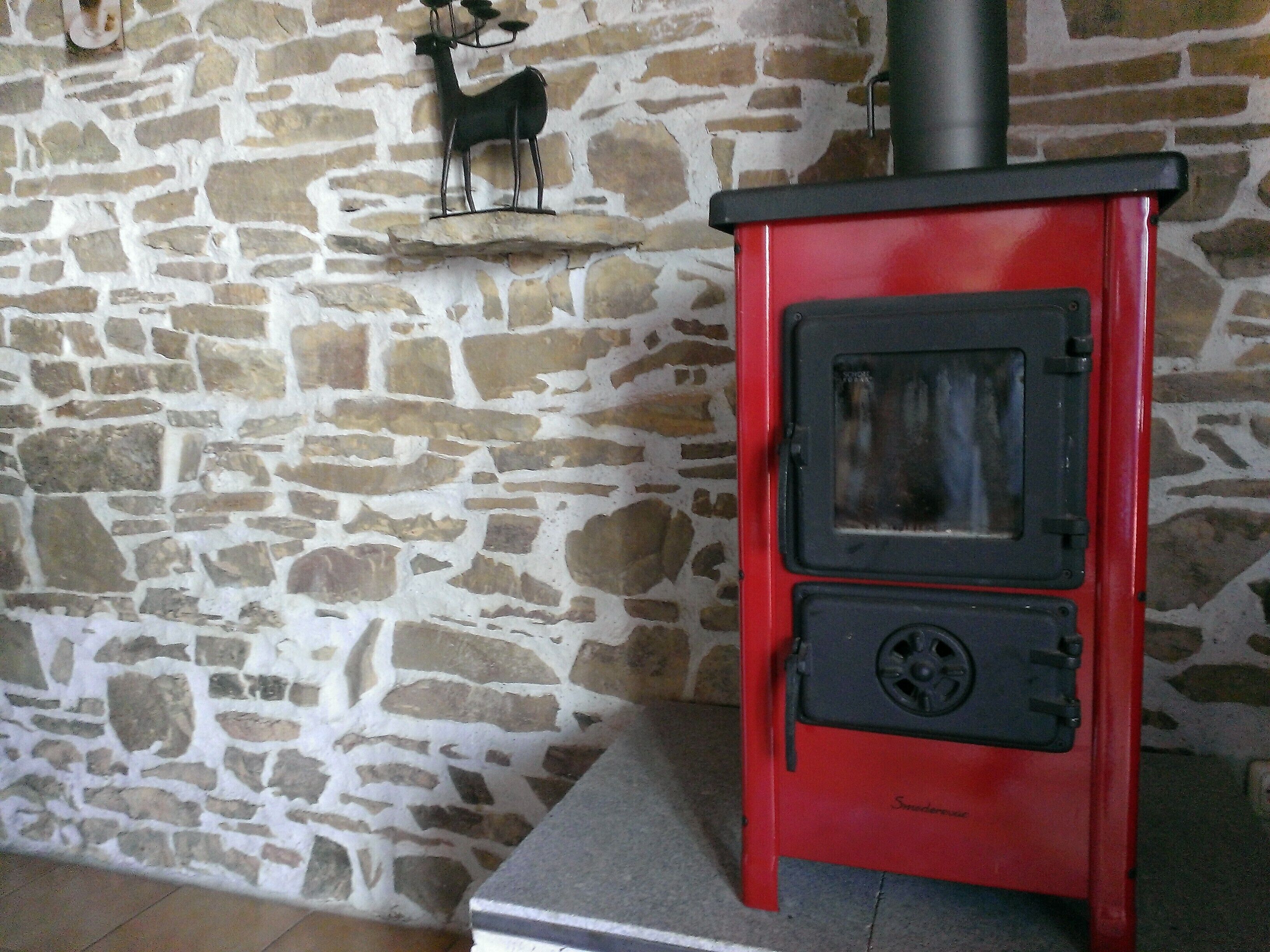 A red wood-burning stove with a chimney, set against a stone wall.
