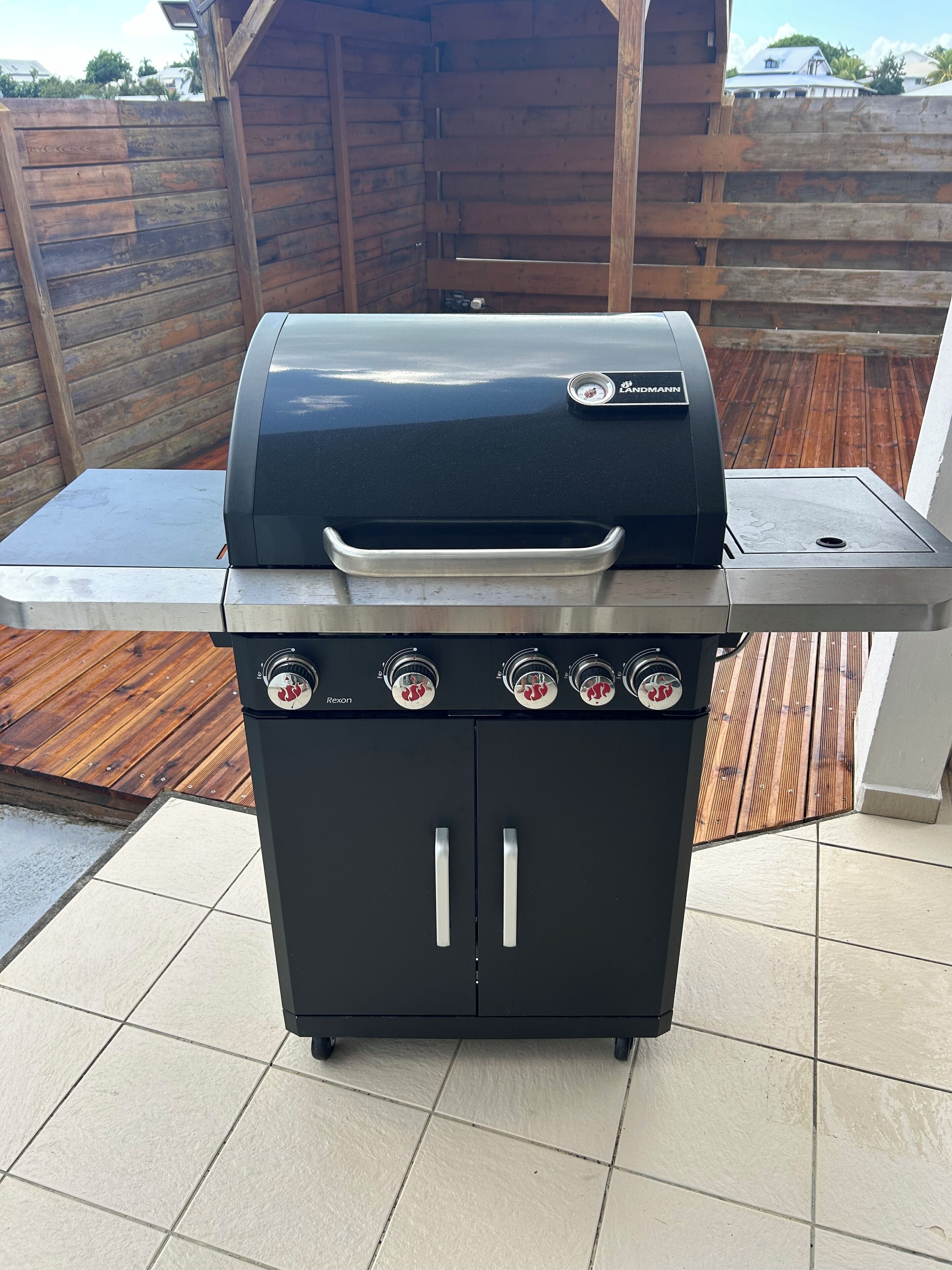 A black barbecue grill with four burners and a side burner, placed on a tiled patio with a wooden deck in the background.