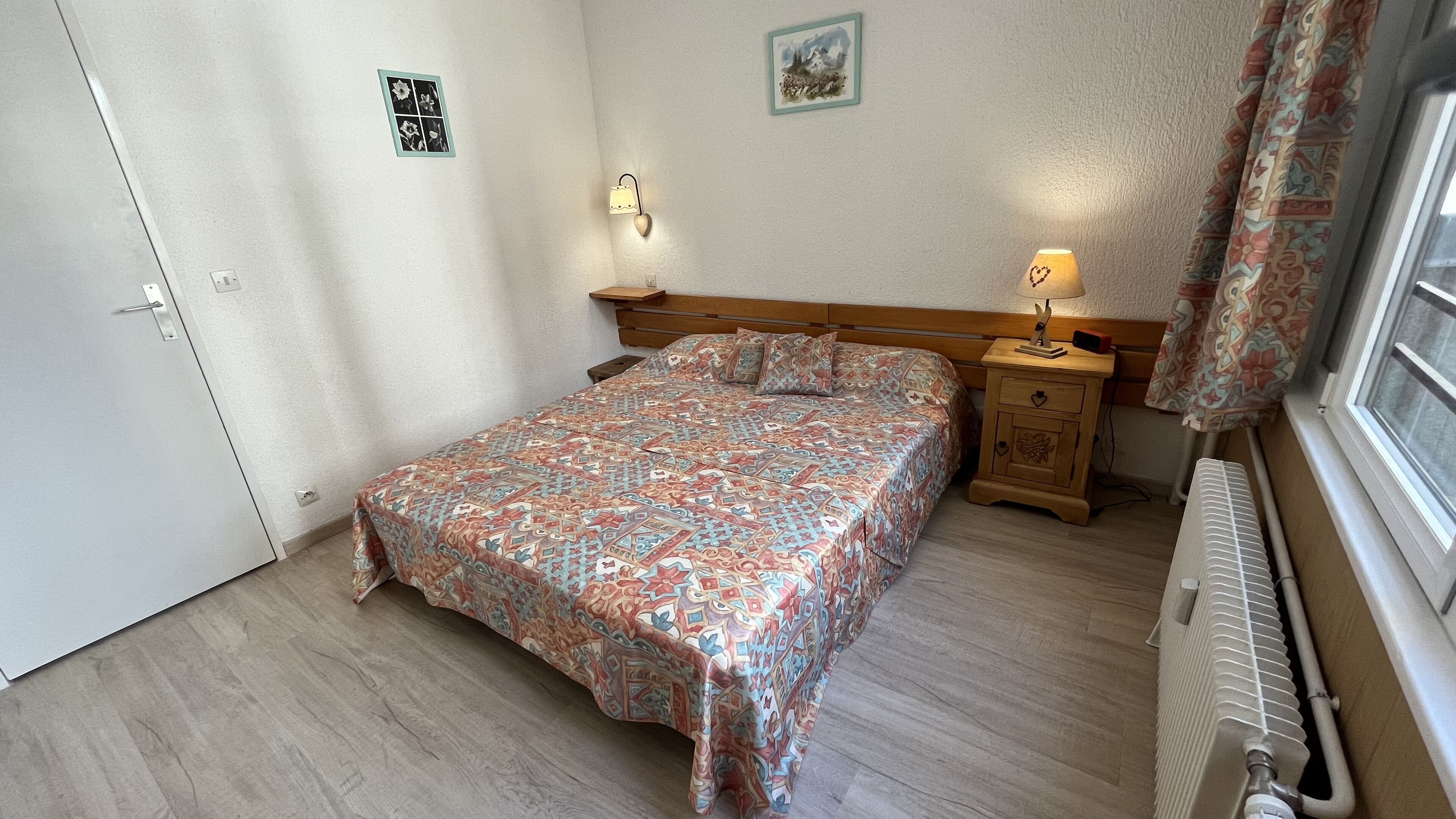 A bedroom with a patterned bedspread, a wooden nightstand with a lamp, a radiator, and a door with a small picture.