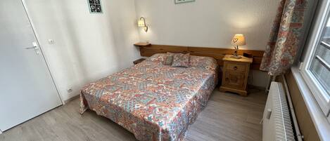 A bedroom with a patterned bedspread, a wooden nightstand with a lamp, a radiator, and a door with a small picture.