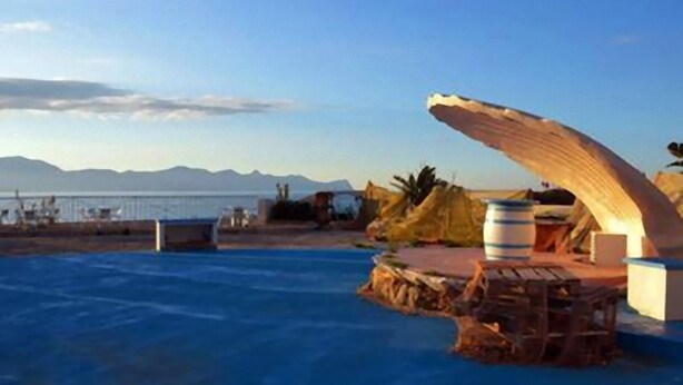 A beachside pool area with a large wooden arch structure, a bar area with stools, and a view of the sea and distant mountains.