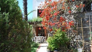 Un chemin mène à un bâtiment blanc, doté d’un mur en pierre et d’un jardin où poussent des fleurs rouges.
