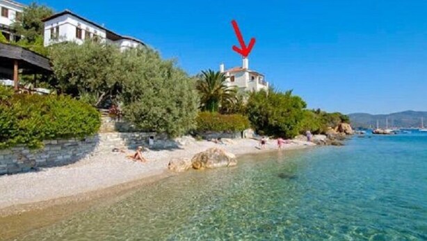 Une propriété située sur la côte, avec une plage de sable fin, un grand rocher dans l’eau et une maison sur laquelle est peinte une flèche rouge pointant vers elle.