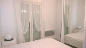 Un dormitorio con una cama, una mesita de noche con lámpara y una ventana con cortinas translúcidas.