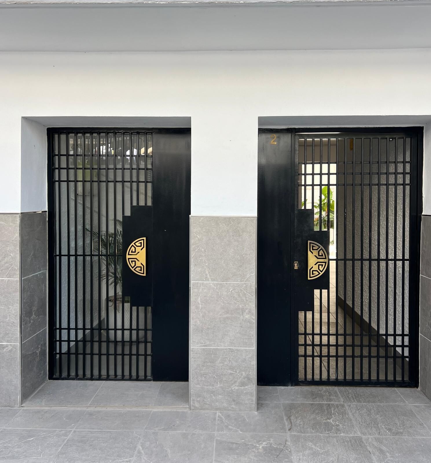Two black metal gates with a number '2' on one and a decorative emblem on the other, set in a modern building with grey stone tiles.
