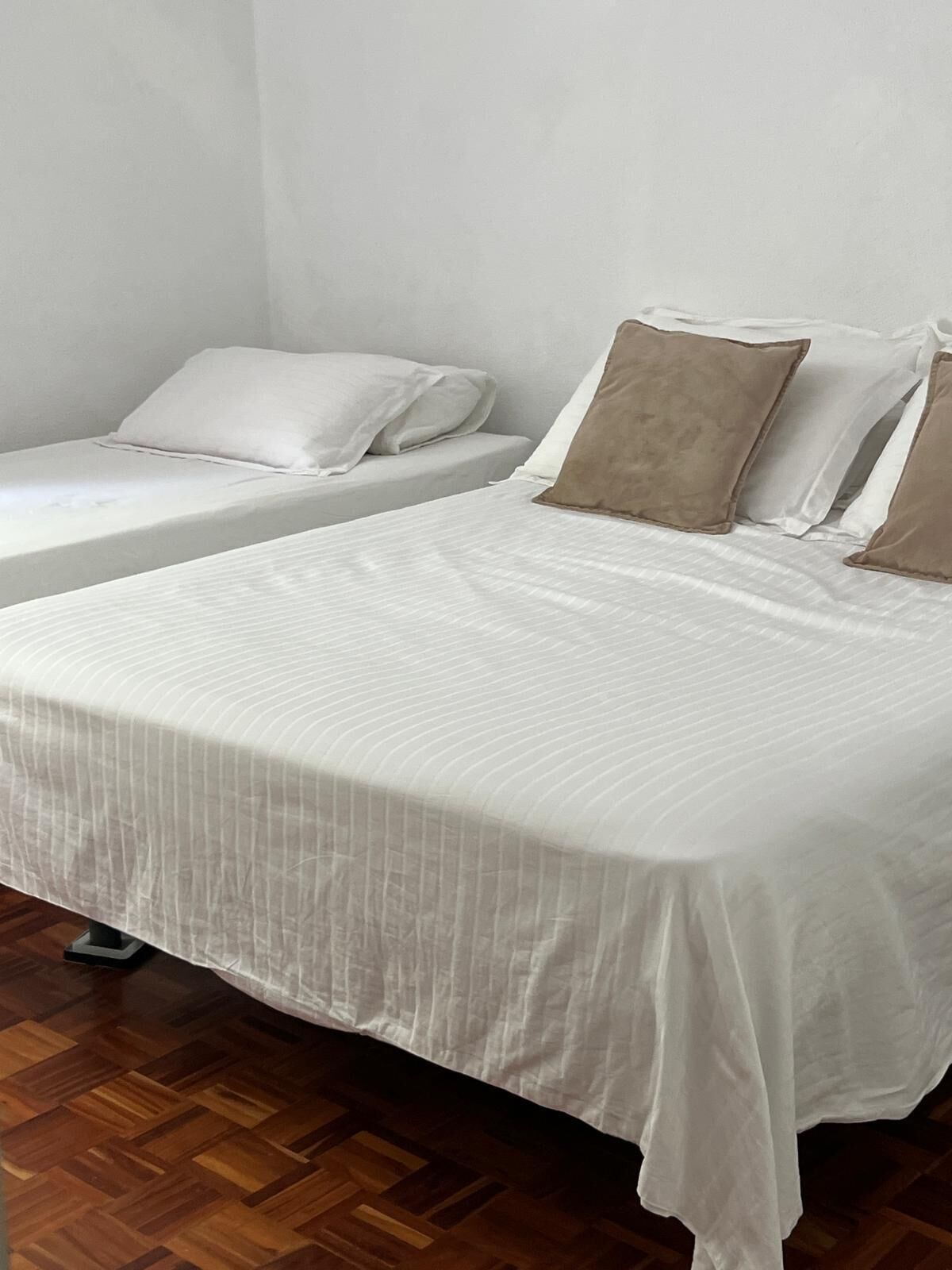 A bedroom with two single beds, each with white bedding and brown pillows. The room has wooden flooring and white walls.