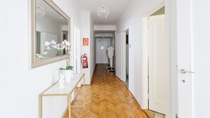 Un couloir doté d’un parquet en bois disposé en chevrons, une console blanche sur laquelle est posée une plante en pot, un grand miroir et une porte sur laquelle est accroché un extincteur.