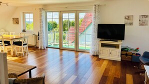 Un vaste salon doté d’un plancher en bois, un coin repas avec une table et des chaises, un téléviseur et une porte-fenêtre coulissante donnant sur un balcon surmonté d’un toit rouge.