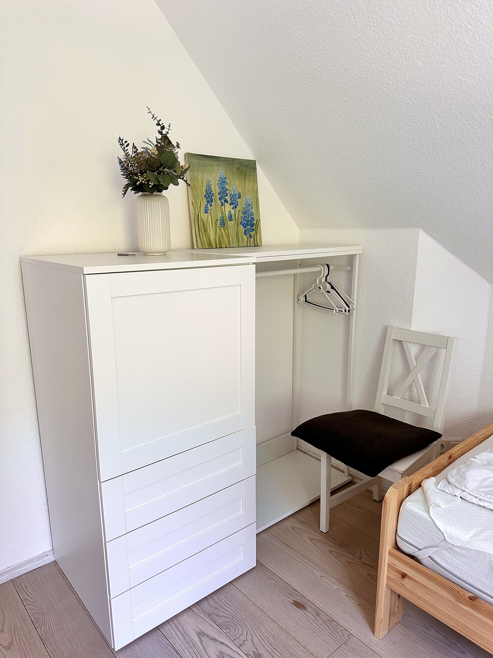 Un bureau blanc avec des tiroirs, une petite chaise, un porte-manteau mural et un tableau représentant des fleurs accroché au mur.