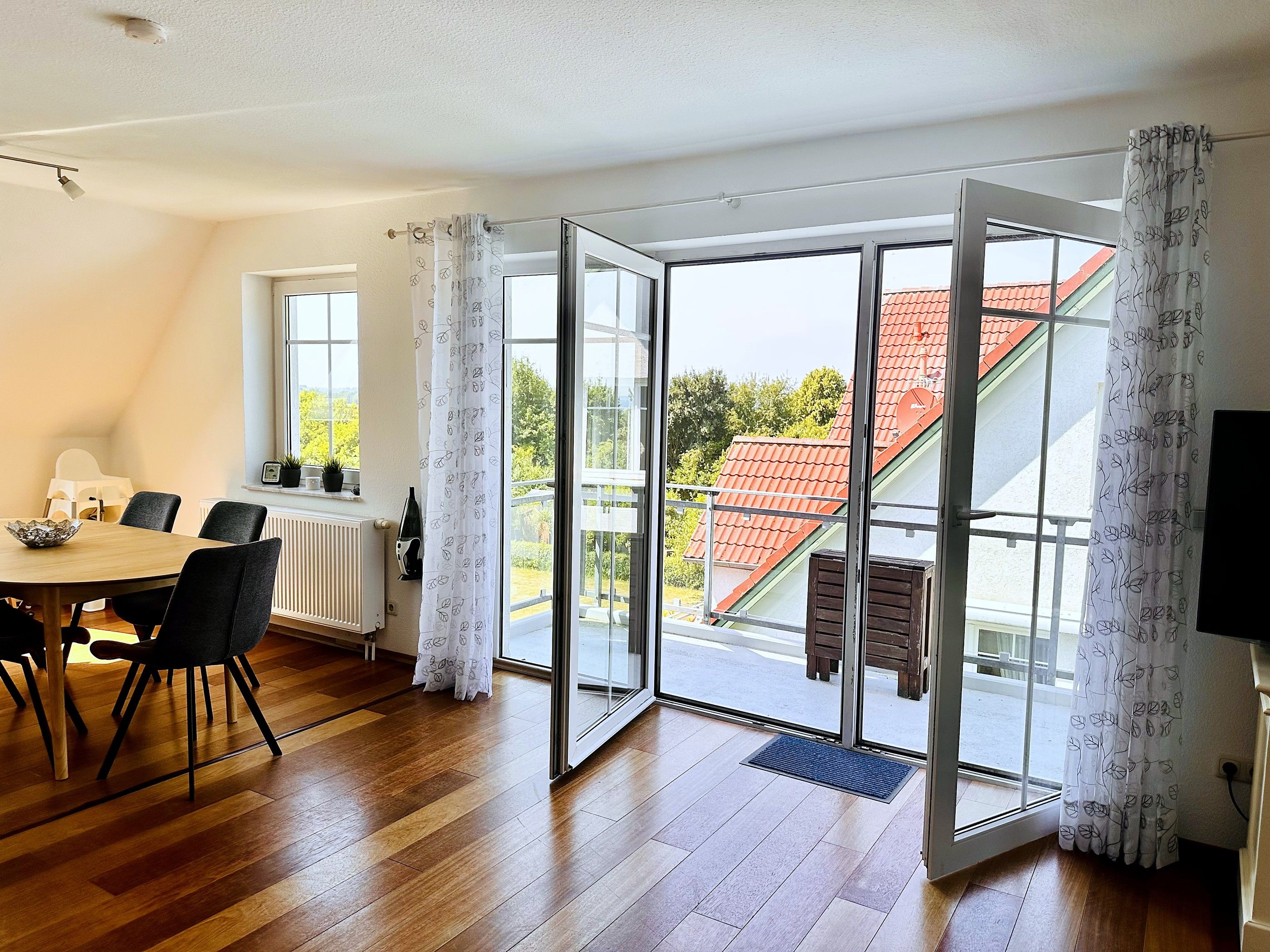 A room with a dining table, chairs, a radiator, a window with a view of a red-roofed building, and a door leading to an outdoor area.