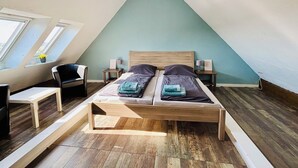 Une chambre avec un plafond en pente, un lit avec deux oreillers, une chaise, une petite table et une table de chevet avec une lampe.