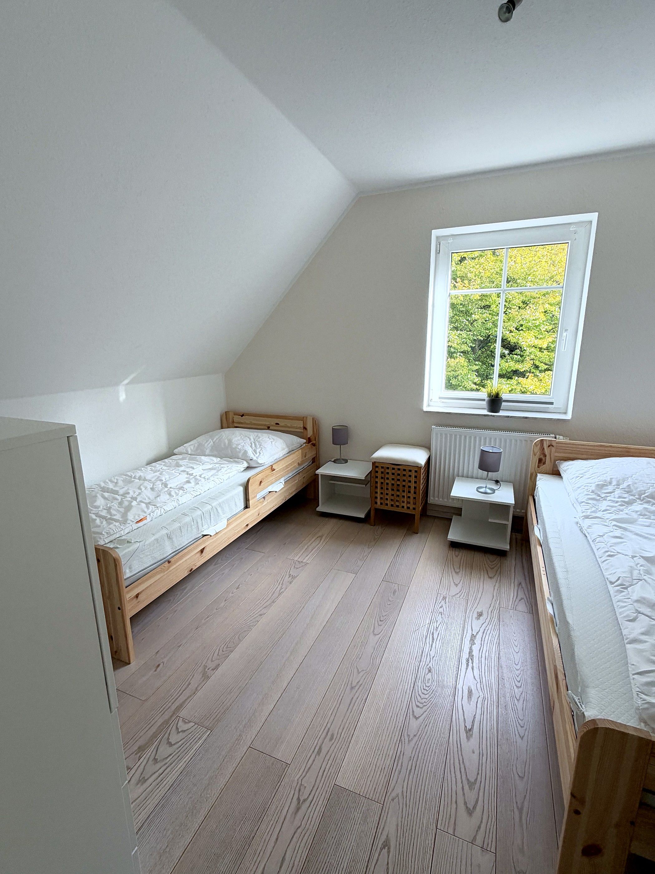 A small, single-room apartment with two wooden beds, a window with a view of greenery, and a radiator.