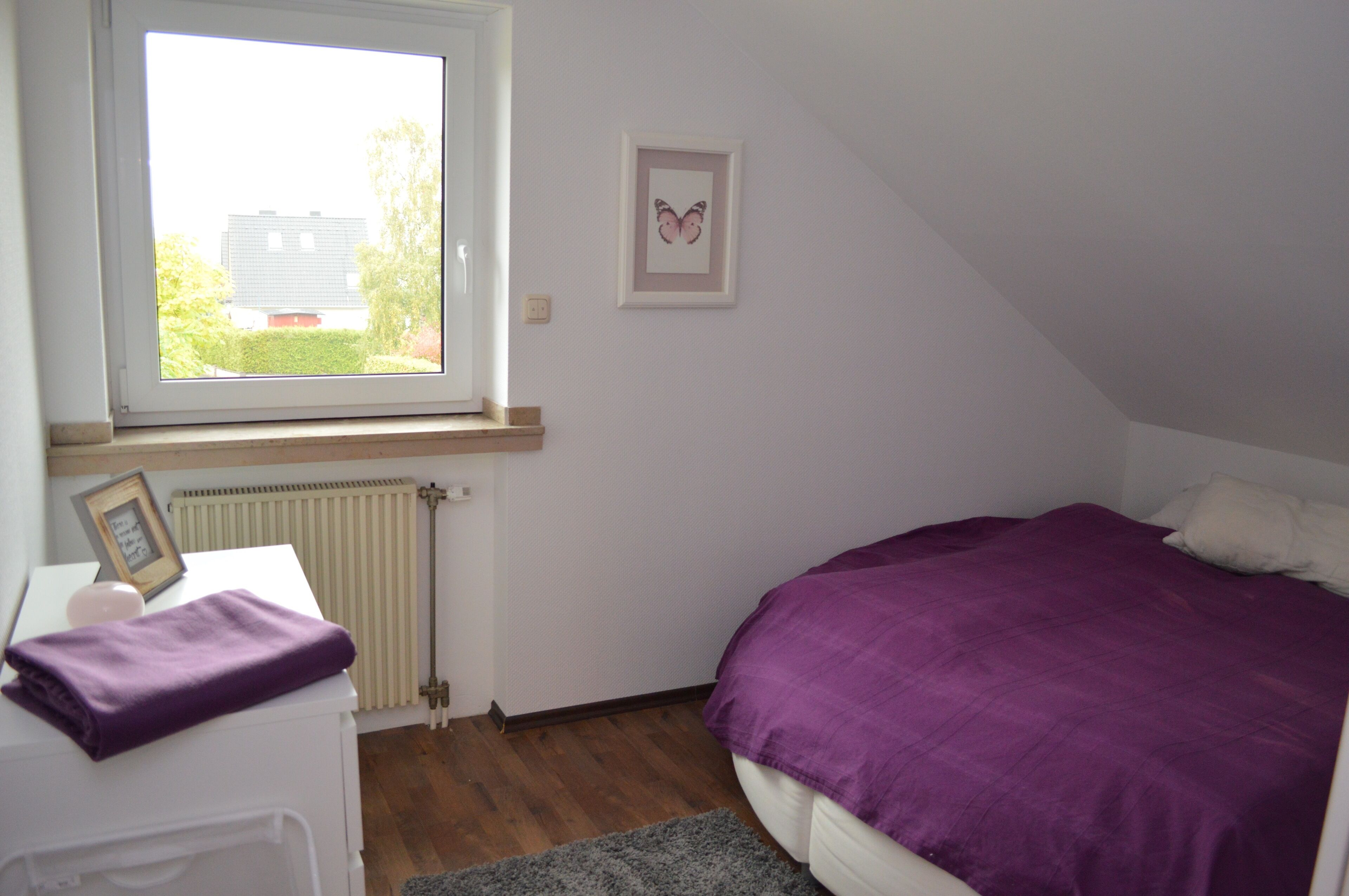 Una camera da letto con una finestra, un letto con coperta viola, un quadro appeso al muro e un termosifone.