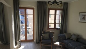 Un salon avec un plancher en bois, un canapé, un fauteuil et une table. Il y a deux fenêtres avec des rideaux, un lustre et un tableau encadré au mur.