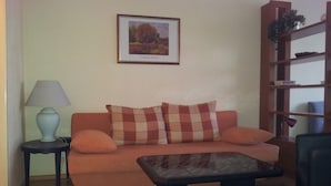 A living room with a sofa, a small table, a lamp, a bookshelf, and a framed picture on the wall. - Apartment City Center 2 (Wien)