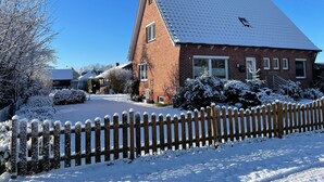 A brick house with a sloped roof, surrounded by snow-covered bushes and a wooden fence.