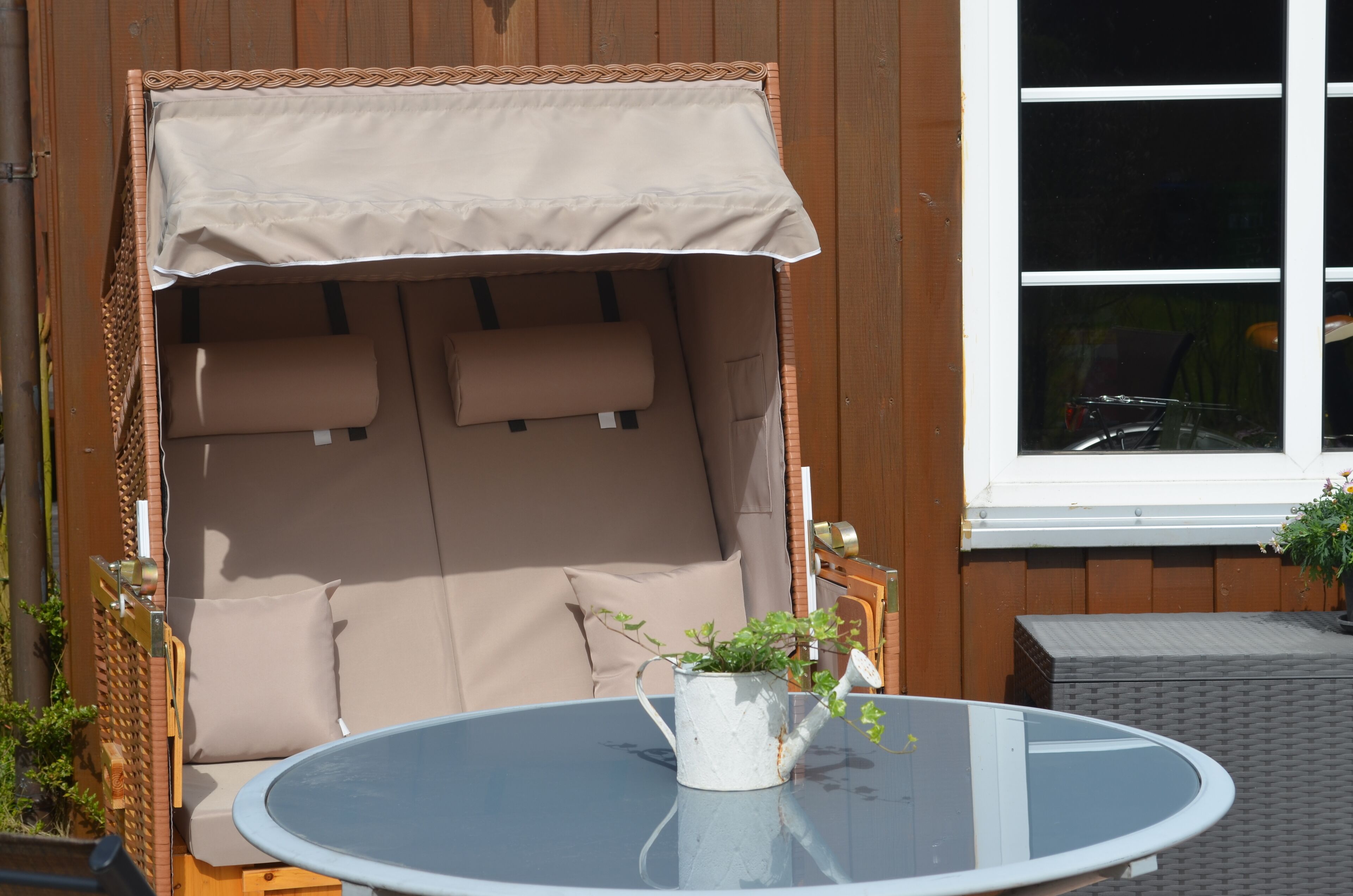 A sunlit outdoor seating area with a cushioned bench, a glass table, and a potted plant.