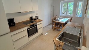 A modern kitchen with white cabinets, a wooden countertop, a built-in oven, and a sink with a wooden tray. There is a dining table with chairs and a window with curtains. - tastefully furnished & quiet apartment in West Lichterfelde (Berlin)