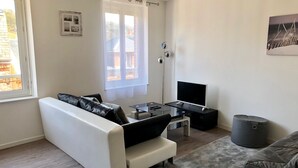 A modern living room with a white sofa, a glass coffee table, a flat-screen TV, and a grey trash bin. - BEAUTIFUL APARTMENT 2 STEPS FROM THE BEACH / NEAR TO THE BEACH! (Étretat)