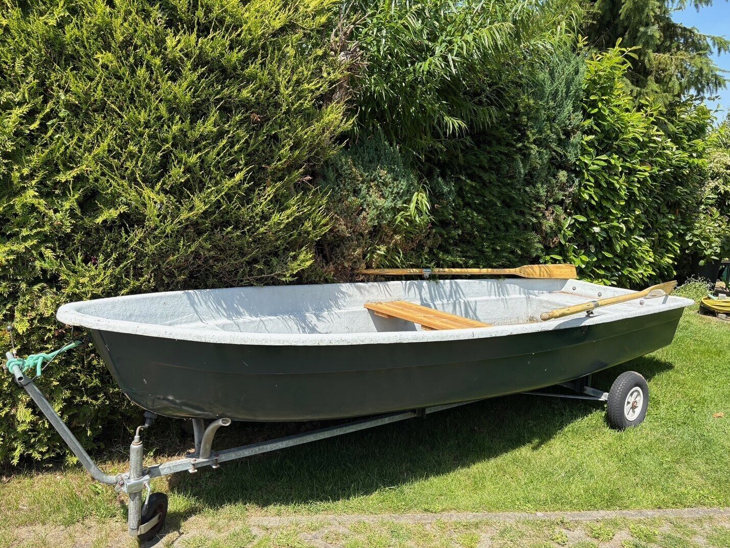 A small, green motorboat with a wooden interior, parked on a trailer in a grassy area with dense green foliage in the background.