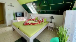 Ein modernes Schlafzimmer mit Schräge, einem Bett mit grüner Bettwäsche, einem Fernseher auf einem Schrank und einem kleinen Nachttisch mit Lampe.