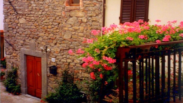 Un bâtiment en pierre avec une porte rouge, une petite fenêtre et un balcon orné de fleurs roses.