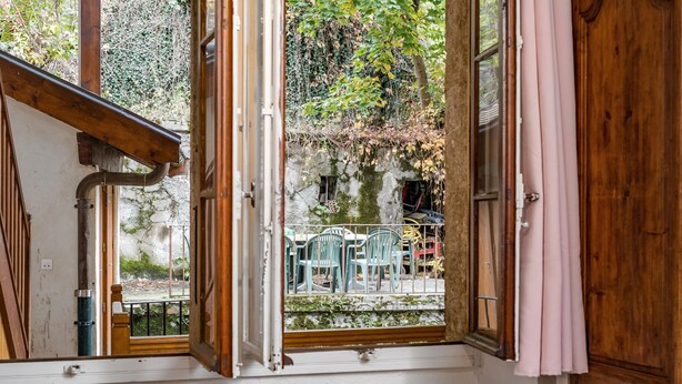 A room with a large window, wooden beams, and a view of outdoor furniture and greenery. - charming studio in the heart of the old town (Annecy)